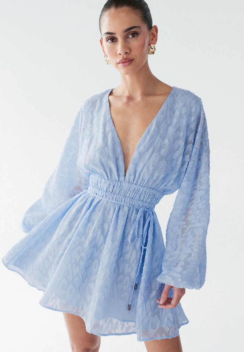 Lichtblauwe jurk van doorschijnende, gestructureerde stof met een diepe V-hals en lange mouwen. Heeft een elastische tailleband en een strikdetail met metallic accenten.