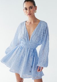 Svetlo modra obleka z v-izrezom in dolgimi rokavi, s teksturno vzorcem in elastičnim pasom. Lahka silhueta v obliki črke A.