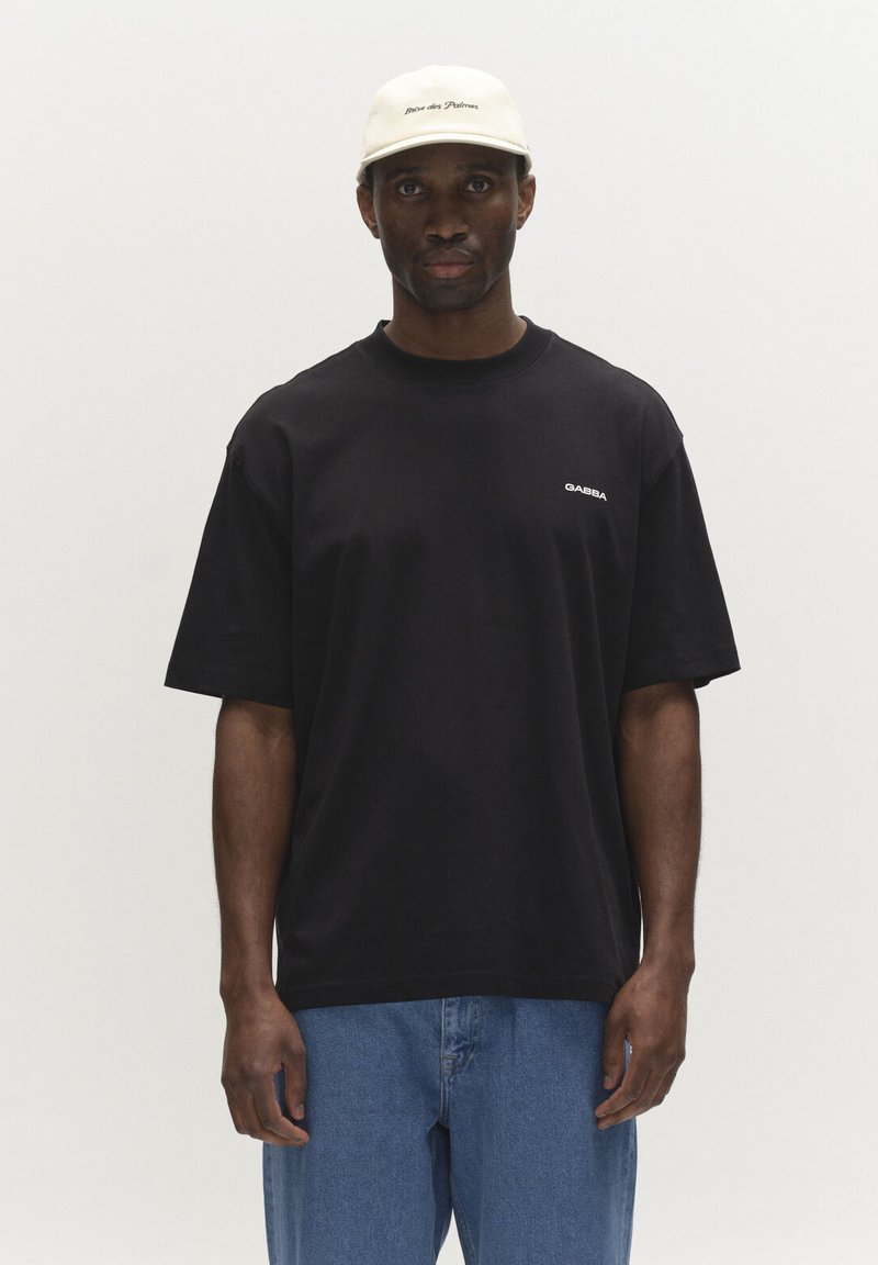 Homme portant un t-shirt Gabba noir, un jean bleu et une casquette blanche avec le texte "Brice des Palmes", debout devant un fond clair uni.