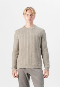 Pull en maille torsadée beige clair avec un col rond, des poignets et un ourlet côtelés. Texture douce, design ajusté, porté avec un pantalon en velours côtelé gris.