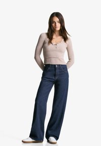 Rangsávos bézs crop top V-kivágással és három gombbal, magasderekú, sötétkék, széles szárú farmerral és fehér sportcipővel párosítva.