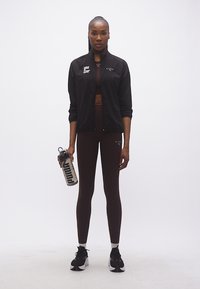 Γυναίκα με μαύρο αθλητικό μπουφάν και κολάν κρατάει ένα μπουκάλι νερού Puma, στέκεται μπροστά σε απλό λευκό φόντο.