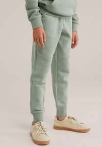 Gaiši zaļi sporta bikses no mīksta auduma, ar elastīgiem piedurknēm un sānu paneļiem, apvienoti ar bēšiem sporta apaviem.