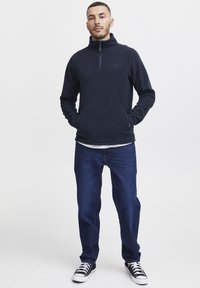 Sudadera de forro polar azul marino con cuello de media cremallera, combinada con jeans azul oscuro y zapatillas negras con cordones blancos. Detalle de marca minimalista.
