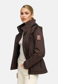 Vrouw met blond haar in een paardenstaart, gekleed in een bruine jas over een beige top en lichte broek, staand tegen een effen witte achtergrond.