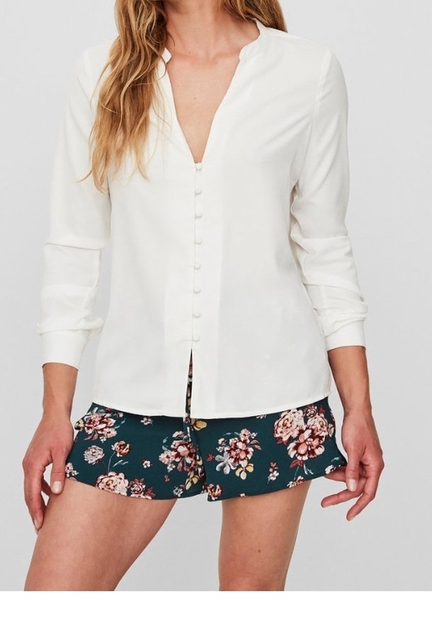 Donna che indossa una camicia bianca a maniche lunghe con bottoni e pantaloni corti verde scuro con motivi floreali rosa e bianchi.