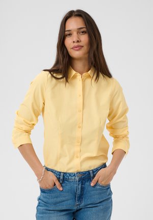 Femme aux longs cheveux bruns portant une chemise jaune à manches longues et un jean bleu, les mains dans les poches, debout devant un fond uni.