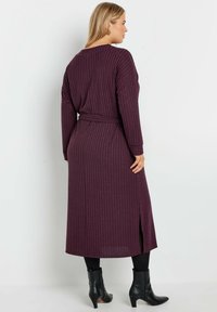 Lång, räfflad klänning i mörklila med en bälte i midjan, utrustad med långa ärmar och slitsar på sidorna, som kombineras med svarta ankelstövlar.