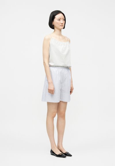 Γυναίκα με κοντά μαύρα μαλλιά, ντυμένη με λευκό αμάνικο τοπ, ανοιχτό μπλε ριγέ σορτς και μαύρες μυτερές μπαλαρίνες, στέκεται μπροστά σε απλό φόντο.