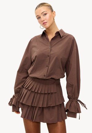 Blouse en coton marron avec fermeture à boutons et mini-jupe plissée. Présente des manches larges avec liens et des volants superposés, créant un design texturé.