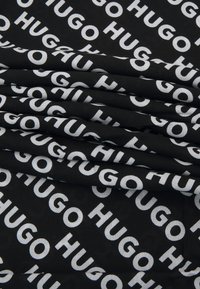 Svart stoff med gjentatt hvit "HUGO" merkevare i ulike orienteringer, med en glatt tekstur og foldet design.