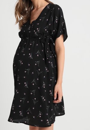 Vrouw die een zwarte bloemen zwangerschapsjurk draagt met korte mouwen en een zijstrik in de taille tegen een effen achtergrond.