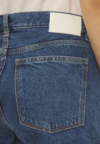 Jeans de mezclilla azul oscuro con costuras naranjas, con un parche de cuero blanco en la cinturilla y un bolsillo trasero visible.