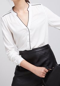 Blouse blanche avec passepoil noir le long du col et de la patte de boutonnage, manches longues avec poignets à boutons, portée avec une jupe noire texturée.