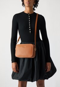 Braune Leder-Umhängetasche mit Stickdetails, rechteckiger Form und goldenen Beschlägen. Getragen zu einem schwarzen, gerippten Langarmkleid und einem Rock.