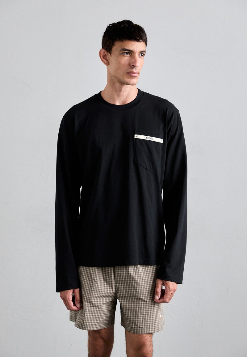 Svart langermet T-skjorte med frontlomme og hvit etikett, kombinert med beige rutete shorts. Jevn stoff, enkelt design.