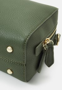 Πράσινη, υφασμένη δερμάτινη τσάντα με κλείσιμο φερμουάρ, χρυσό υλικό και διακοσμητικές σπουδές στο μπροστινό μέρος. Συμπαγές ορθογώνιο σχήμα.