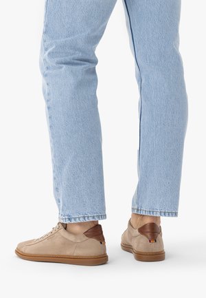 Donji dijelovi nogu u svijetlo plavim trapericama i bež semiš tenisicama s detaljima smeđe kože na peti, na bijeloj pozadini.