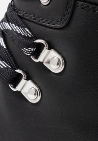 Bota de cuero negro con cordones a rayas blancas, herrajes plateados y una textura suave. Primer plano de los detalles de los cordones y ojales.