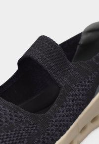 Μαύρο πλεκτό παπούτσι χωρίς κορδόνια με φαρδιά ελαστική λωρίδα στο πάνω μέρος και ανοιχτό μπεζ υφή σόλα.