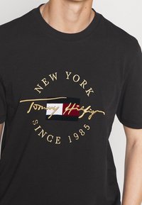 Černé tričko s vyšitým zlatým textem "New York Since 1985" a logem Tommy Hilfiger s červenou, bílou a námořnickou modrou vlajkou.