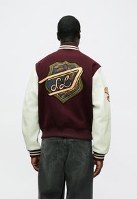 Bordo spalvos universitetinio stiliaus striukė su baltomis odinėmis rankovėmis, nugarinėje dalyje – išsamus siuvinėtas skydelio dizainas. Ribos apykaklė ir rankogaliai.