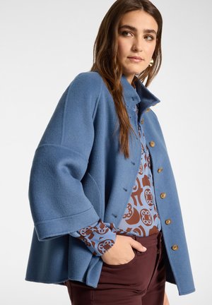 Femme portant un manteau bleu boutonné par-dessus un chemisier à motifs et un pantalon bordeaux, la main dans la poche, regardant légèrement sur le côté.