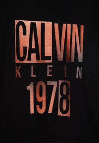 Svart genser med stor grafisk print i kobbertoner, som viser "CALVIN KLEIN 1978" i fet, moderne typografi.