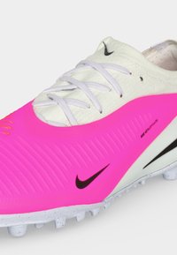 Άσπρο και έντονο ροζ ποδοσφαιρικό παπούτσι Nike με λευκά κορδόνια, μαύρα λογότυπα Swoosh και υφή στη σόλα για παιχνίδι σε συνθετικό χλοοτάπητα.