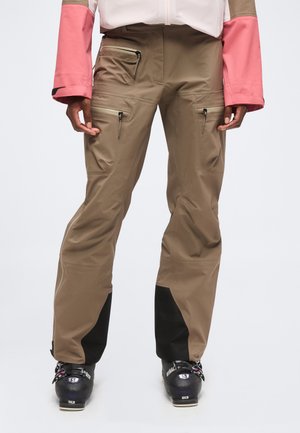 Personne portant un pantalon de ski marron avec des poches zippées et des chaussures de ski noires, debout devant un fond uni.