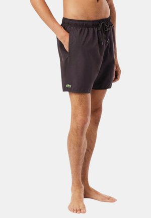 Persona con gambe nude che indossa pantaloncini da bagno neri con tasche laterali e un piccolo logo verde sulla gamba sinistra, in piedi su sfondo bianco.
