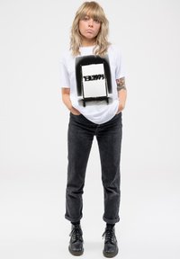 White short-sleeve t-shirt featuring a black graphic design with text, paired with dark grey trousers and black lace-up boots.