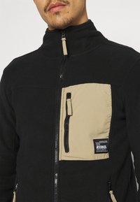 Mężczyzna w czarnej kurtce zapinanej na zamek błyskawiczny z beżową kieszenią na klatce piersiowej z pionowym zamkiem błyskawicznym i metką "The Rebel Project".