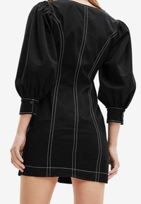 Robe noire avec des manches bouffantes, des coutures blanches contrastantes, une coupe ajustée et des poignets boutonnés avec un ourlet droit.