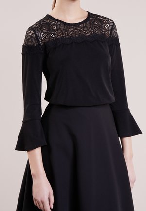 Woman wearing a black dress with lace detail on the upper chest and shoulders and flared three-quarter-length sleeves.