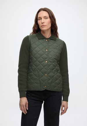 Femme portant une veste matelassée vert foncé avec fermeture par boutons et manches tricotées, debout devant un fond clair uni.
