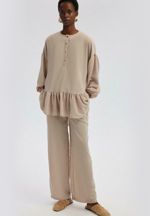 Beige oversized top with button placket and ruffled hem, paired with matching wide-leg pants. Smooth texture, relaxed fit, and minimal design.