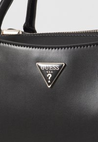 Sac à main synthétique noir avec une texture lisse, présentant un badge logo triangulaire en ton doré avec "GUESS" et "?". Fermeture zippée en haut.