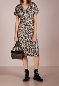 Leopardenmuster-Kleid in Beige und Braun, mit Wickeldesign, kurzen Ärmeln und seitlichen Taschen. Kombiniert mit einer braunen Lackhandtasche und schwarzen Stiefeln.