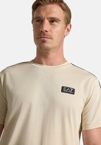 Beige bomulls t-shirt med kort ärm, rundad halsringning, med en svart EA7-logotyp påsydd och svarta kontrasterande detaljer på axlarna.