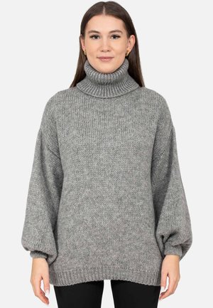 Junge Frau mit langen Haaren, die einen grauen, gestrickten Rollkragenpullover mit Ballonärmeln und schwarze Hose trägt, vor weißem Hintergrund stehend.