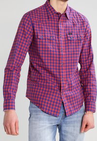 Mann trägt ein rot-blau kariertes Hemd mit Brusttaschen und hellblaue Jeans, steht vor einem schlichten Hintergrund.