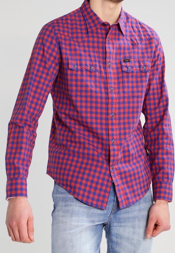 Mann trägt ein rot-blau kariertes Hemd mit Brusttaschen und hellblaue Jeans, steht vor einem schlichten Hintergrund.