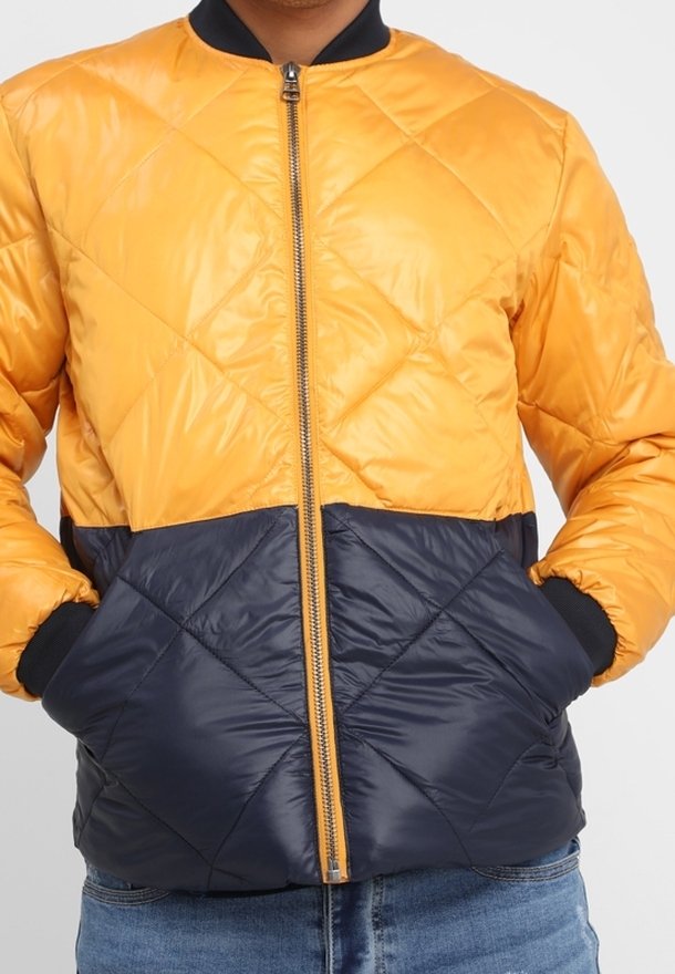 Man in een gele en marineblauwe gewatteerde zip-up jasje met handen in de zakken, gecombineerd met blauwe jeans.