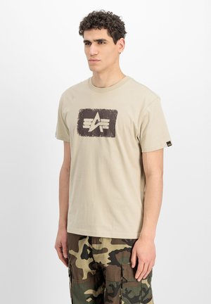 Junger Mann mit lockigen Haaren, der ein beiges Grafik-T-Shirt und Camouflage-Cargohosen trägt, steht vor einem schlichten weißen Hintergrund.