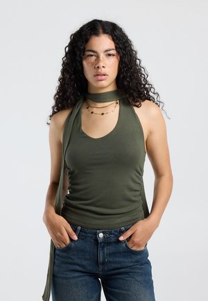 Vrouw met lang krullend haar draagt een olijfgroene mouwloze top, blauwe jeans, gelaagde kettingen, handen in de zakken, neutrale achtergrond.