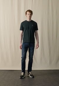 Dark green short-sleeve t-shirt, blue denim jeans, and black sneakers with white accents, standing against a neutral backdrop.