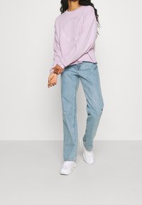 Kvinna i ljuslila sweatshirt med cirkeldesign, ljusblå jeans och vita sneakers som står mot en enfärgad bakgrund.