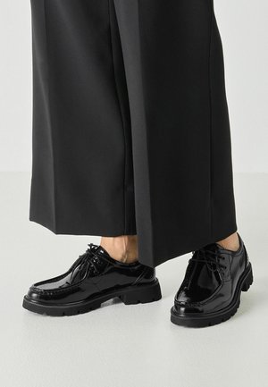 Sorte skinnende læder snørestøvler båret med brede sorte bukser, vist fra midt på læggen og ned på en lys gulvbaggrund.