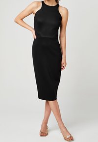 Robe noire midi avec une silhouette ajustée, design sans manches, encolure haute et tissu lisse. Portée avec des escarpins dorés à lanières.